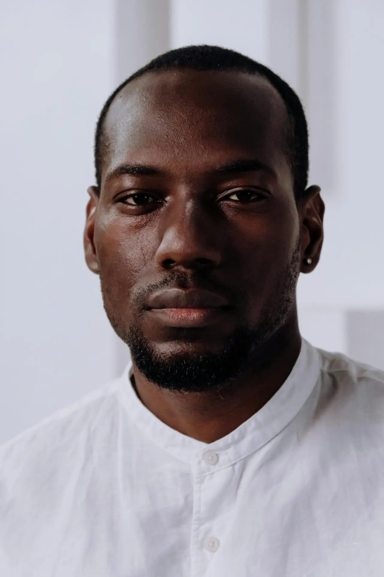 Headshot of a serious black man in a white shirt, indoors with a neutral background.